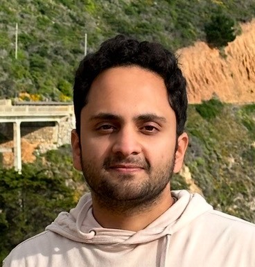 A person in a light hoodie standing outdoors with a scenic background of greenery, a bridge, and rocky terrain.
