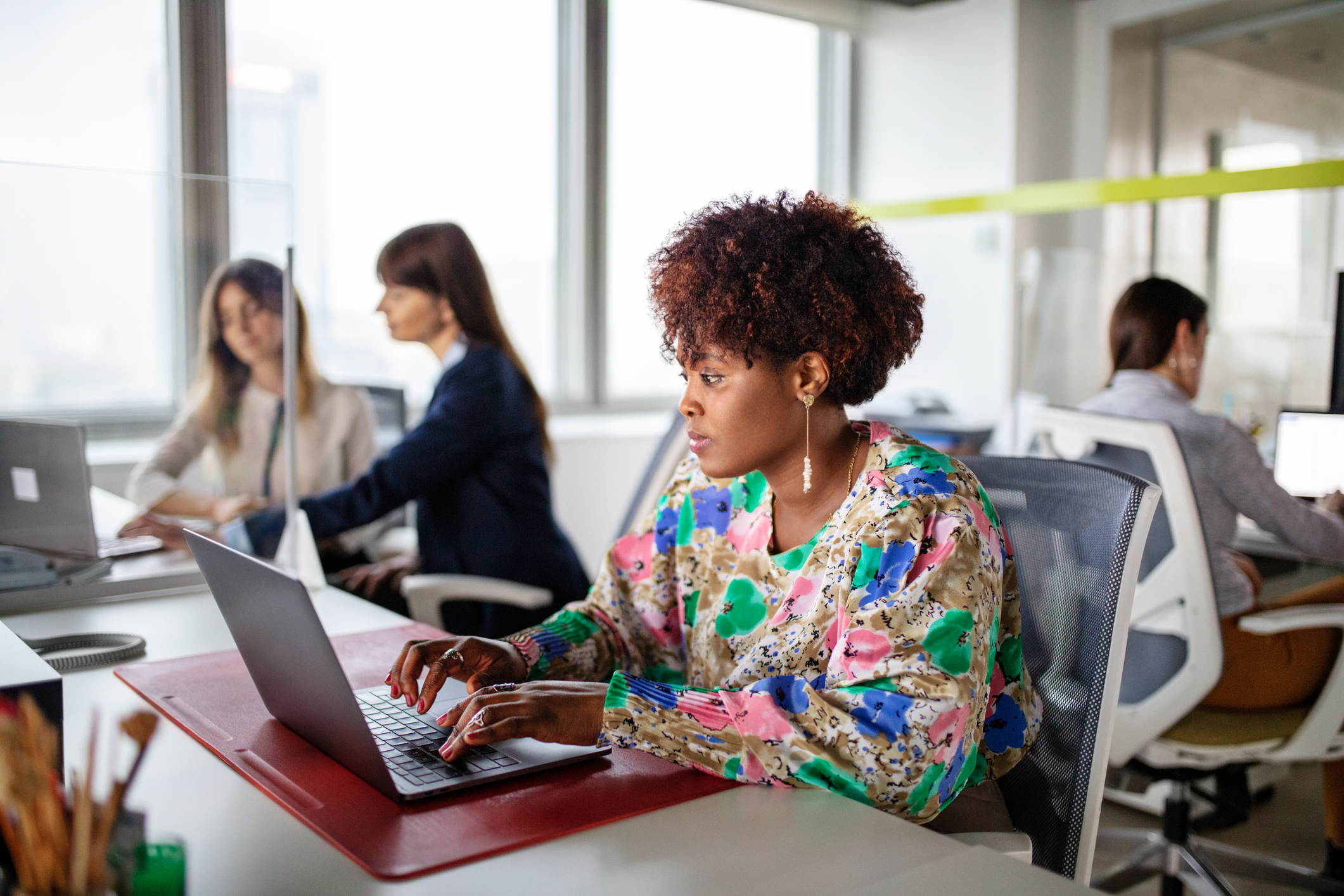 African woman working on laptop while sitting at her desk with colleagues working in background at home.