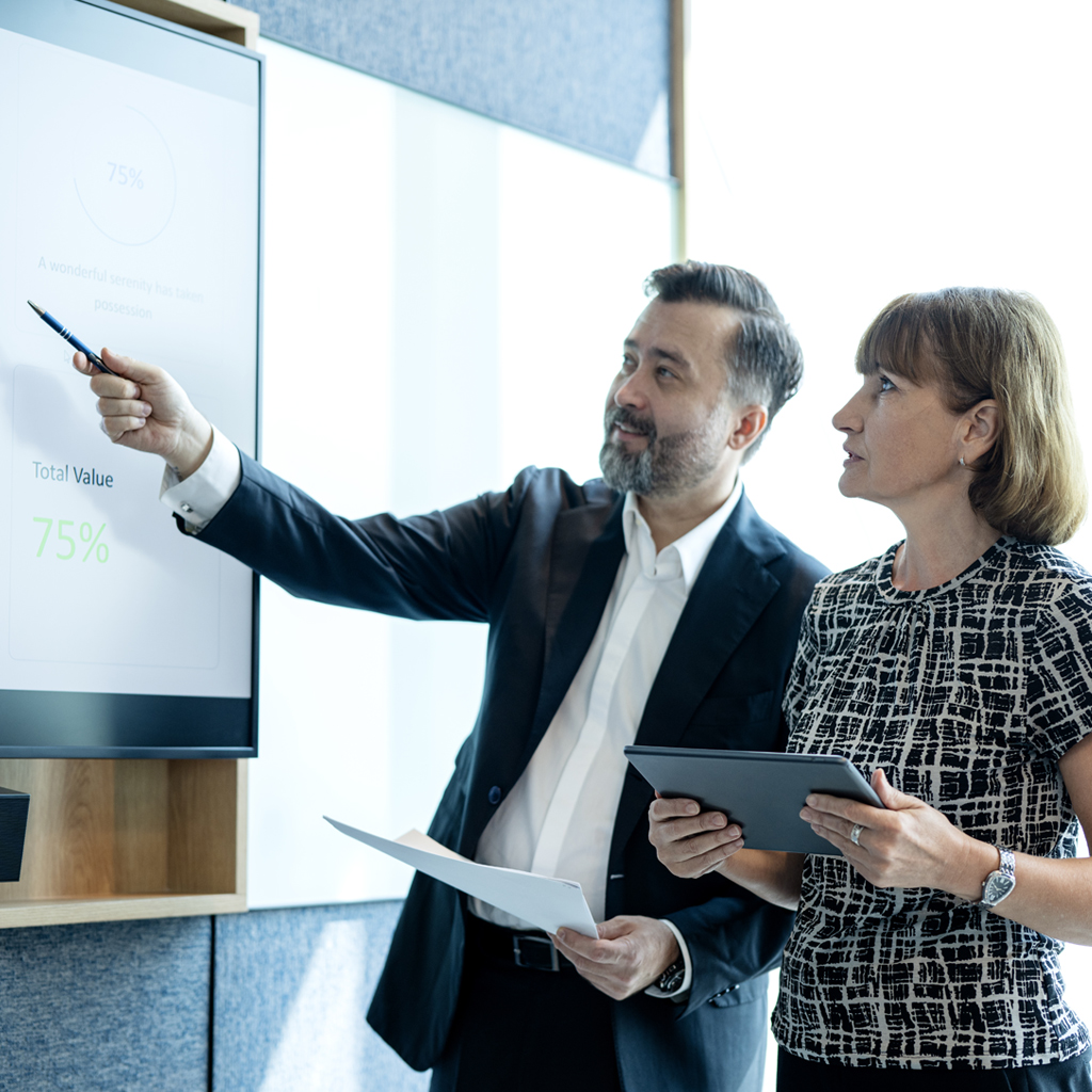 A person in a suit pointing at a presentation screen displaying "Total Value 75%" while another person holds a tablet.