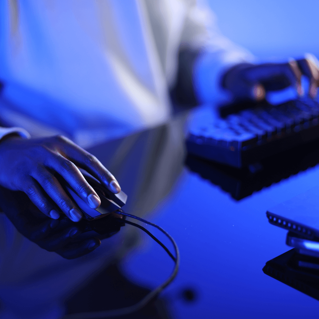 Close-up of a person’s hand using a computer mouse, with a keyboard and reflective surface under blue lighting.