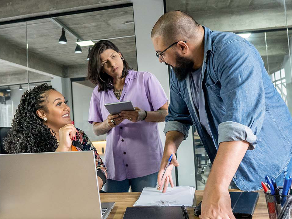 Three colleagues collaborating in a modern office, gathered around a table with a laptop, notebooks, and pens during a product team meeting.