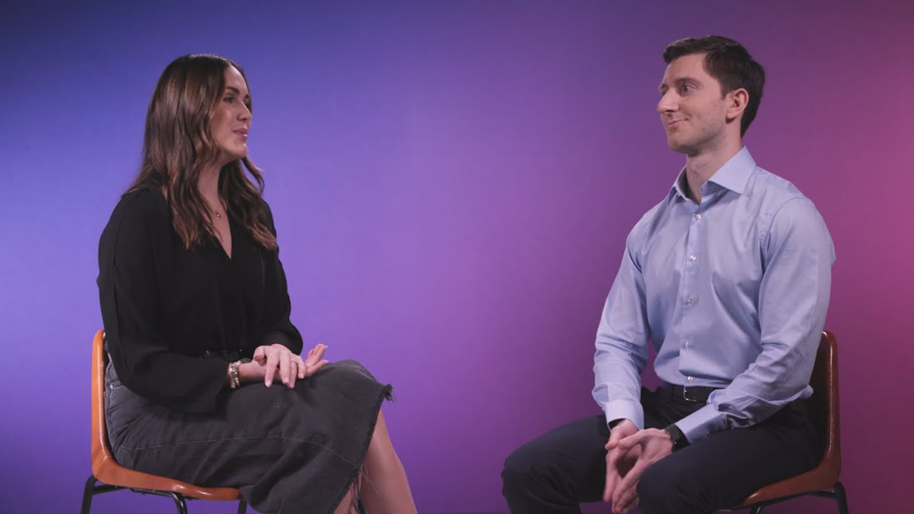 A woman and a man are seated on chairs facing each other, engaged in conversation against a purple gradient background.