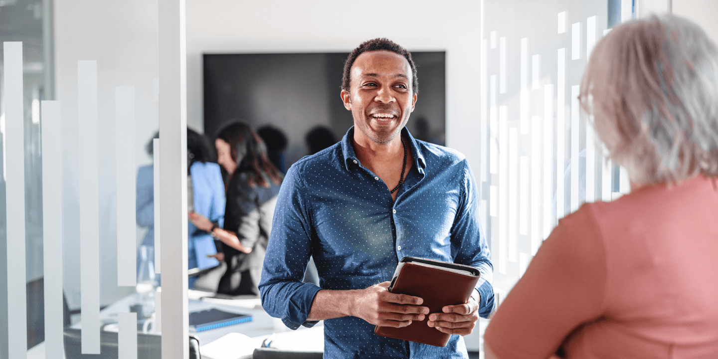 A man is smiling and holding a tablet while talking to a colleague in an office meeting setting, with other people visible in the background.