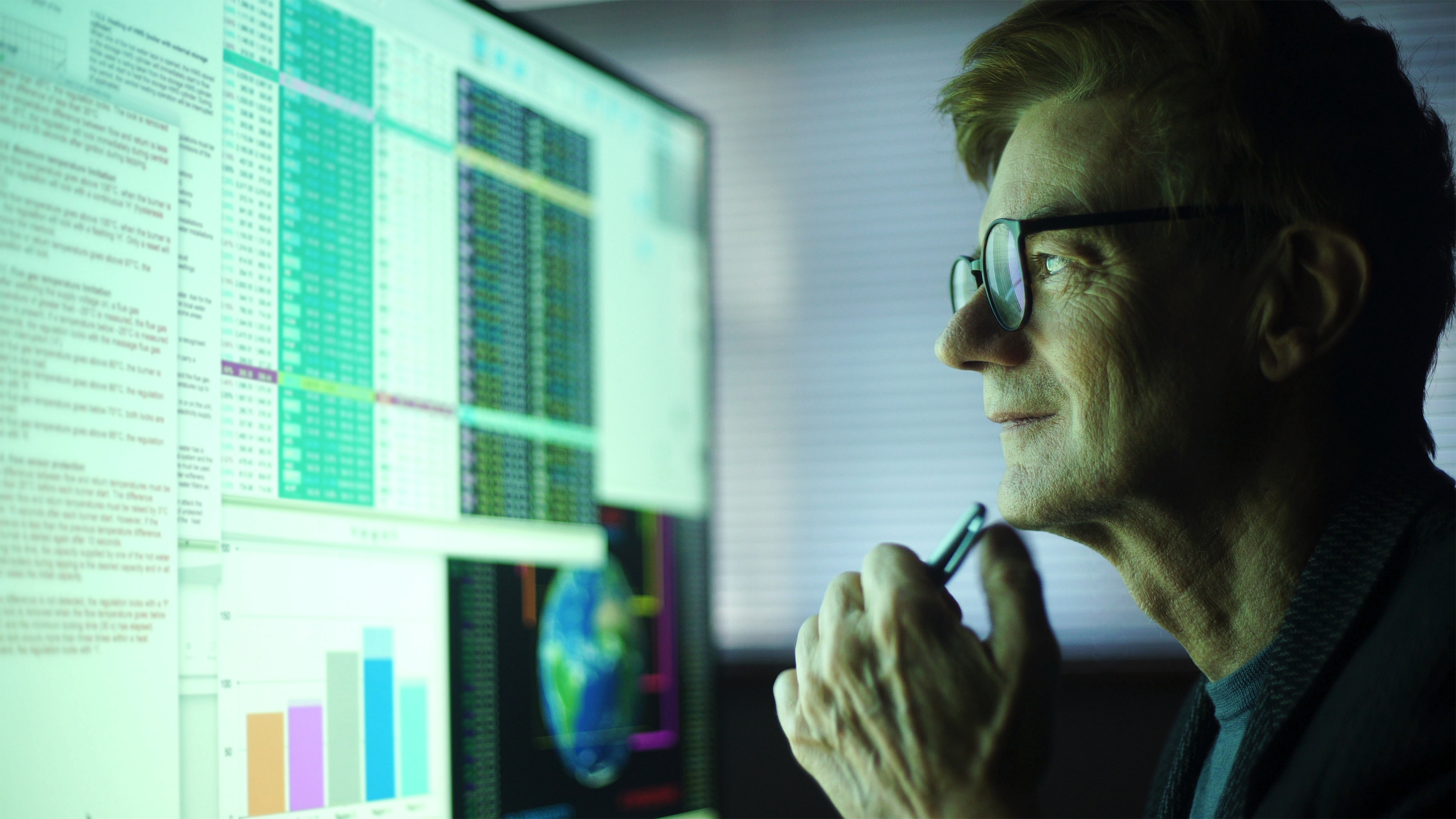 Stock photo of a mature man working in his home office, studying a large computer monitor displaying a variety of numerical data, global information & text.