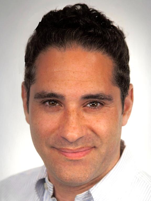 A person with curly dark hair wearing a collared shirt against a plain background.