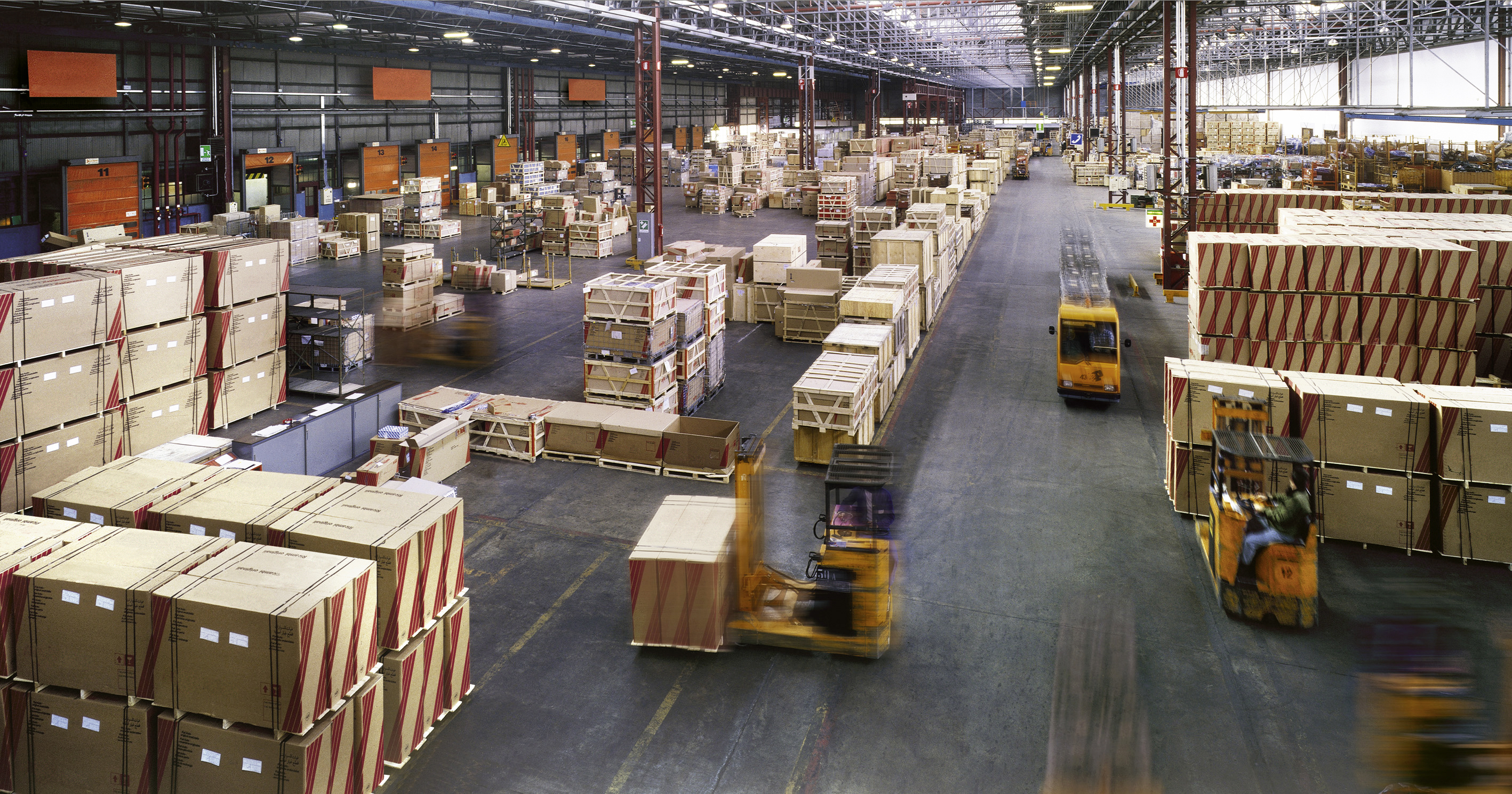 A spacious industrial warehouse filled with stacked pallets and large crates, with several forklifts actively moving materials throughout the facility.