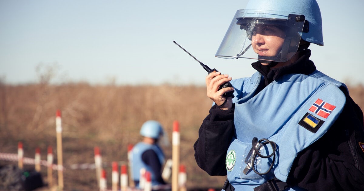 A person wearing protective gear and a blue helmet holds a radio in a field marked with red and white stakes.