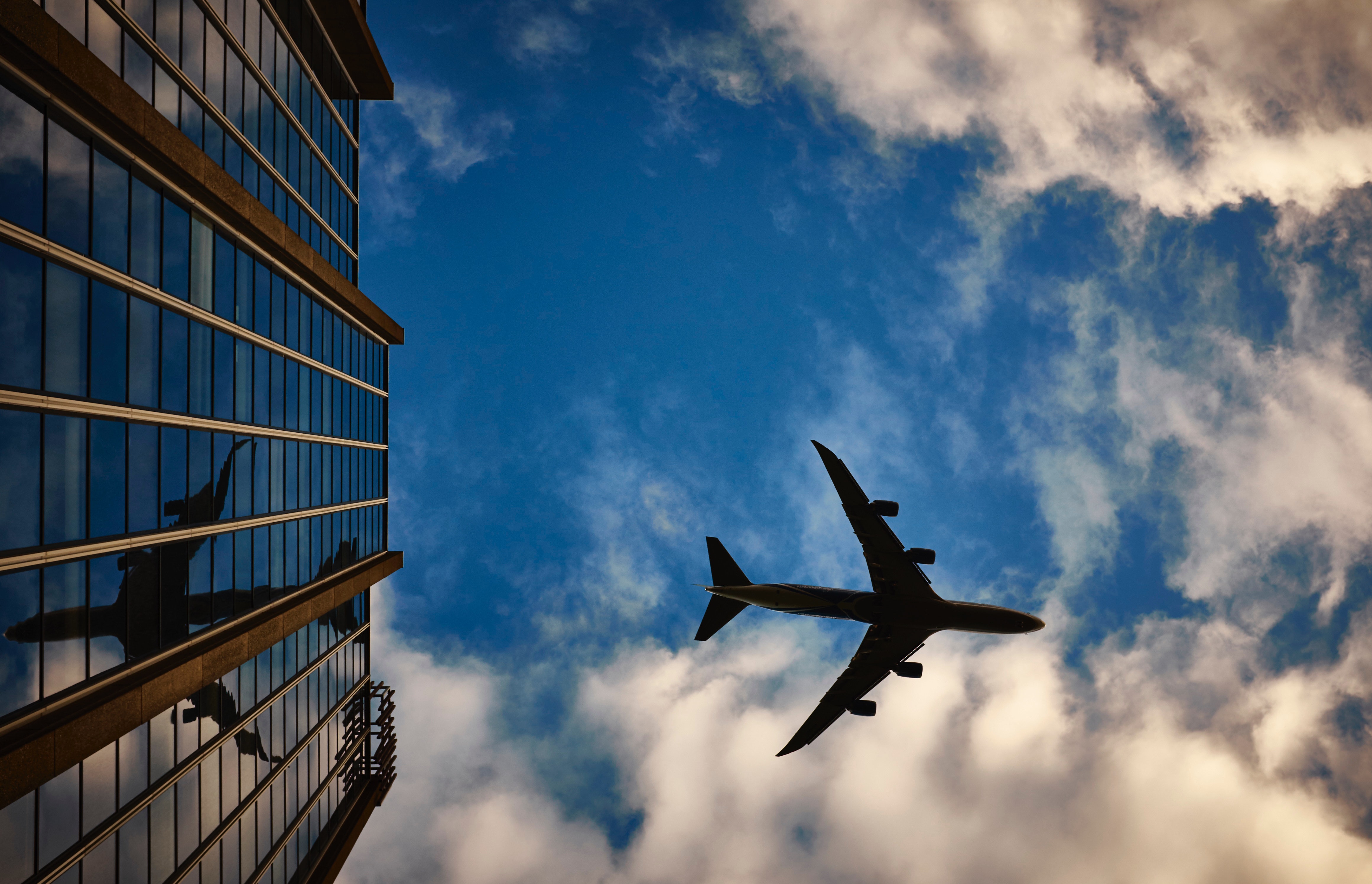 A silhouette of an airplane flying in the sky with clouds, with its reflection visible on the glass windows of a tall building.