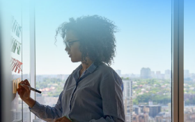 A businesswoman is writing on a glass window with a cityscape visible in the background.