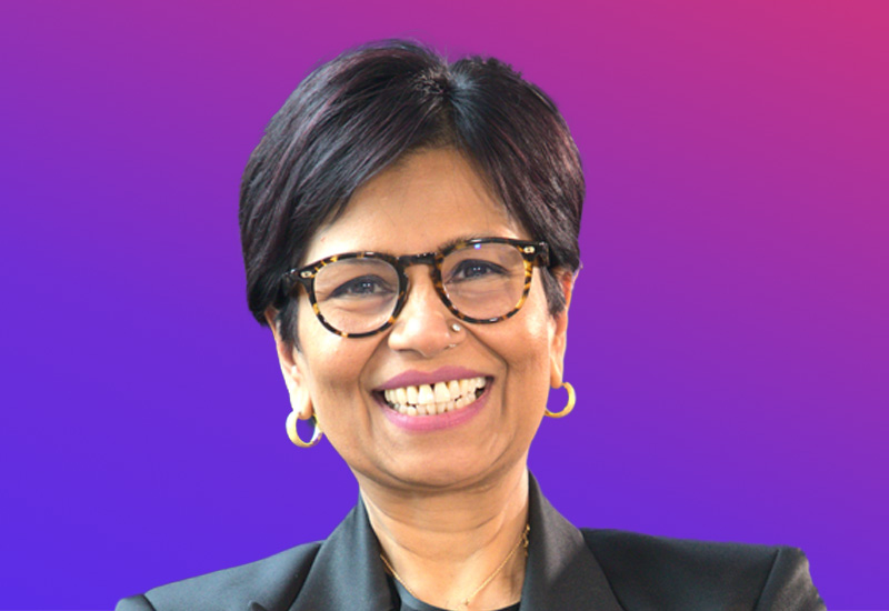 A person with short black hair wearing gold hoop earrings and a black blazer, set against a gradient purple and pink background.