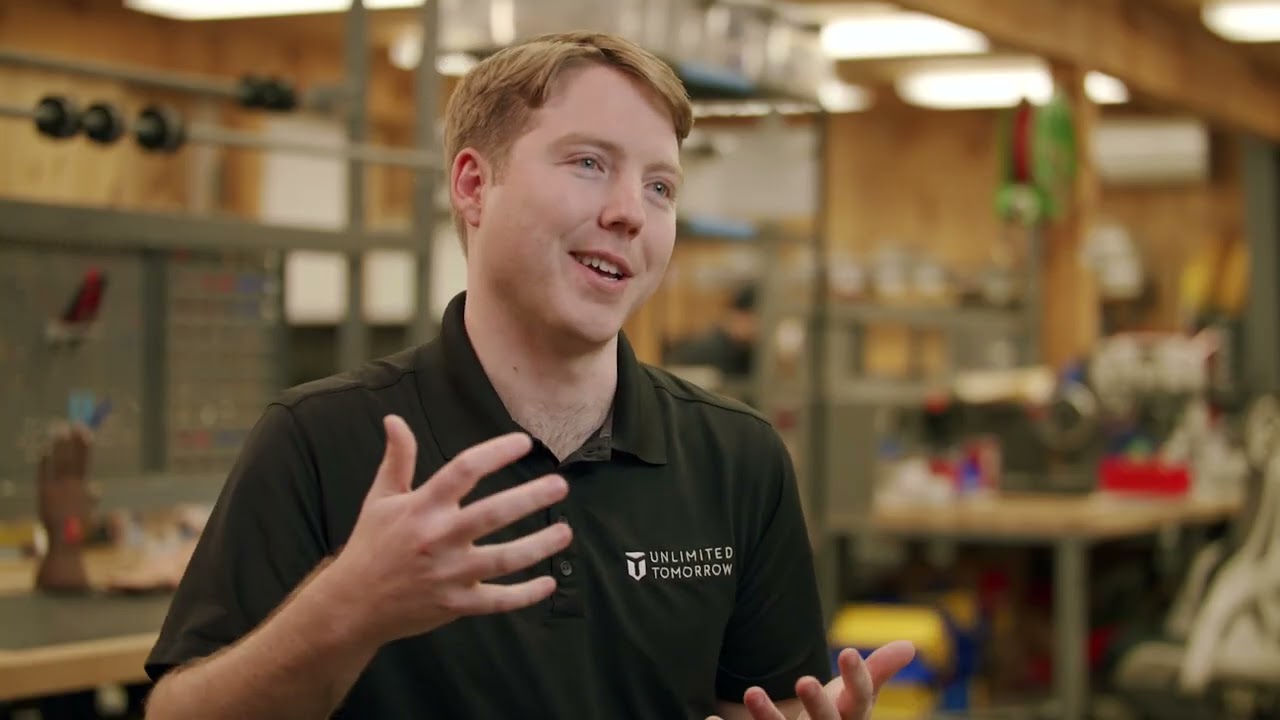 A person wearing an 'Unlimited Tomorrow' logo shirt gestures while speaking in a workshop or lab environment, featuring various tools and equipment in the background.