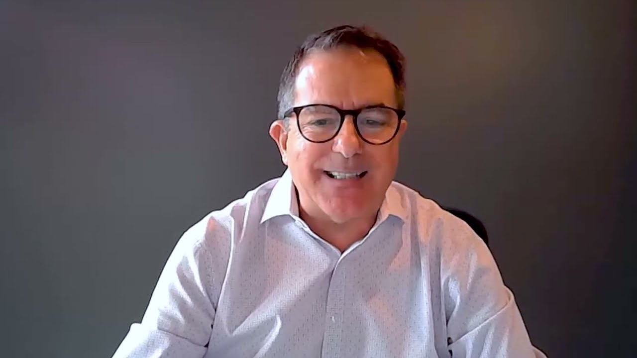 A man wearing glasses and a white shirt is smiling while seated in front of a plain gray background.