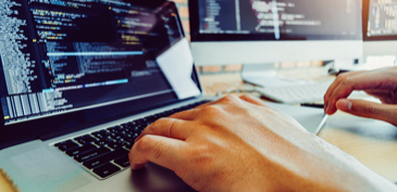 A close-up view of a person coding on a laptop with multiple external monitors displaying programming code, representing software development and coding environments.