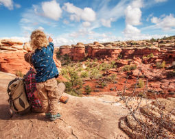 A young child points out into a scenic red rock canyon while hiking, under a bright sky with scattered clouds.
