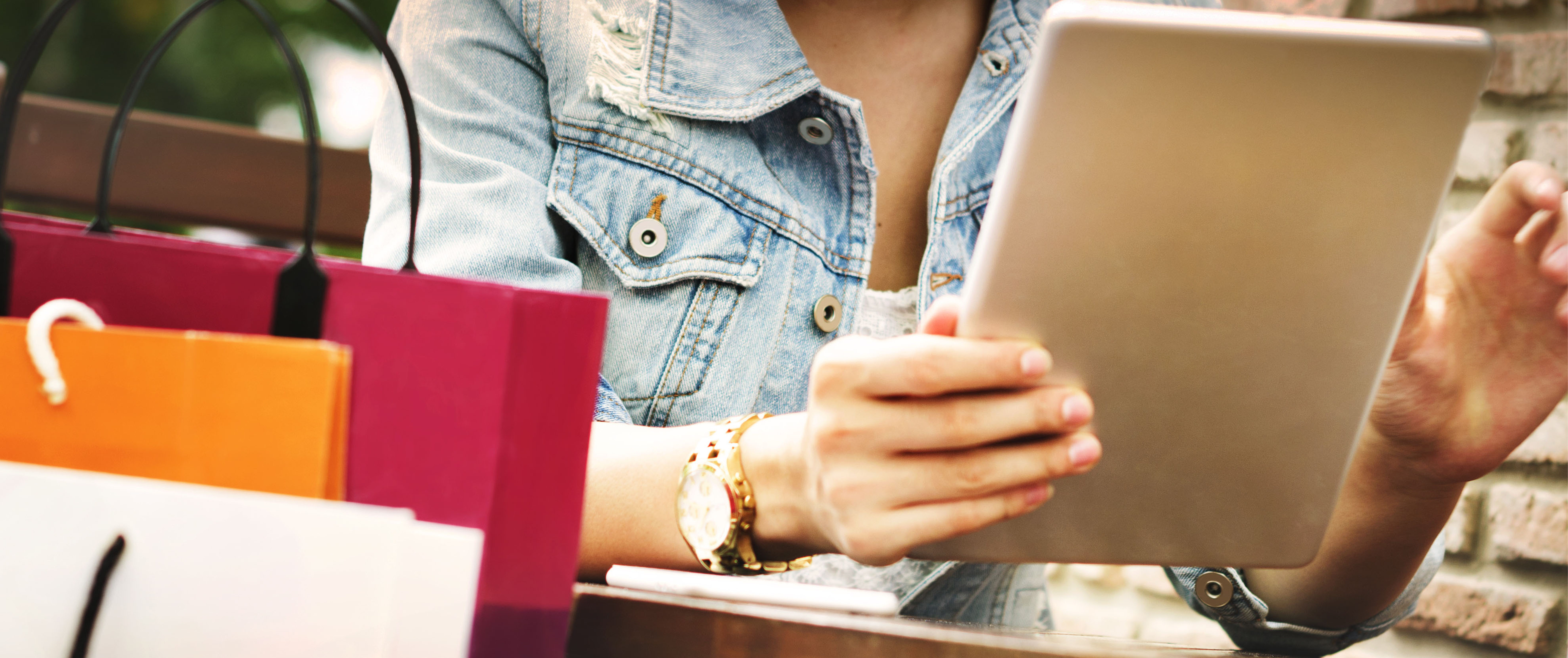 A woman sitting at a table using a tablet for online shopping, with colorful shopping bags beside her.
