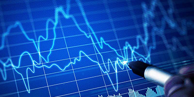 A close-up image of a hand holding a pen, drawing over a digital financial chart with fluctuating blue graph lines, representing trading and investing concepts.