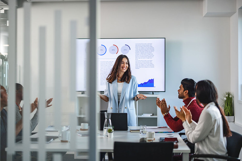 A group of people in a conference room are engaged in a meeting while a presenter stands in front of a screen, smiling as the team applauds. The atmosphere is collaborative and positive, illustrating teamwork and success during a business presentation.