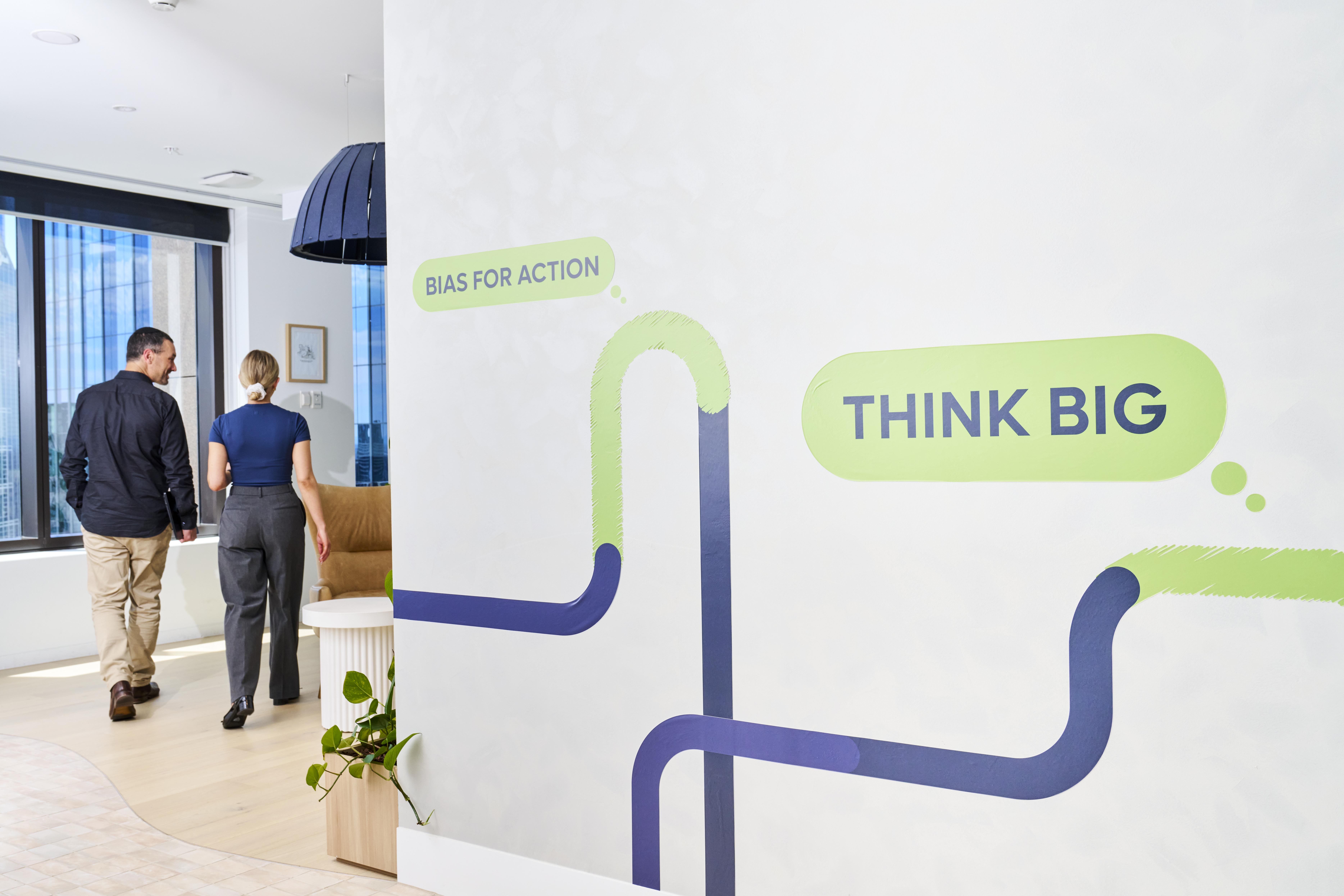 A modern office space at Judo Bank featuring a wall mural with the phrases 'THINK BIG' and 'BIAS FOR ACTION.' Two people are walking through the office, and the mural is a prominent visual element.
