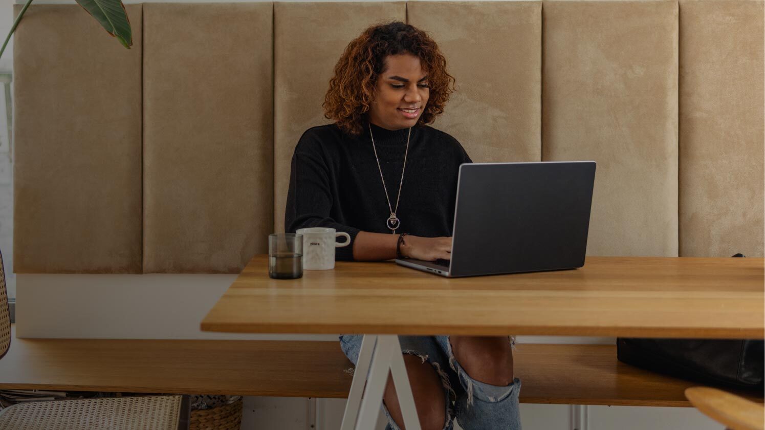 A person sitting at a wooden table with a laptop, a coffee mug, and a glass, against a beige cushioned wall.