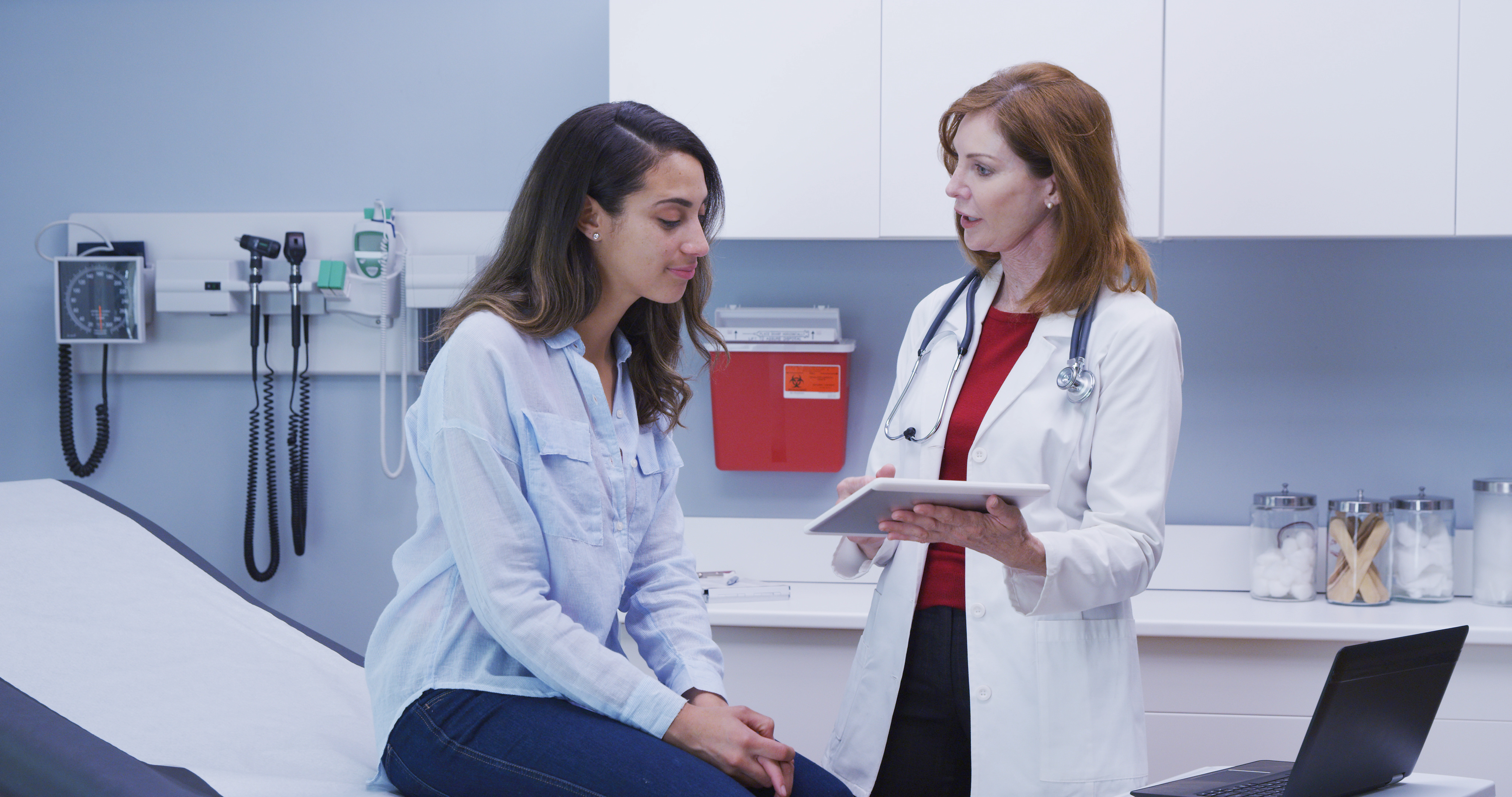 A female doctor is consulting with a young female patient in a modern clinic exam room. The doctor is holding a digital tablet and discussing healthcare with the patient, who is seated on the examination table. Medical equipment is visible in the background.