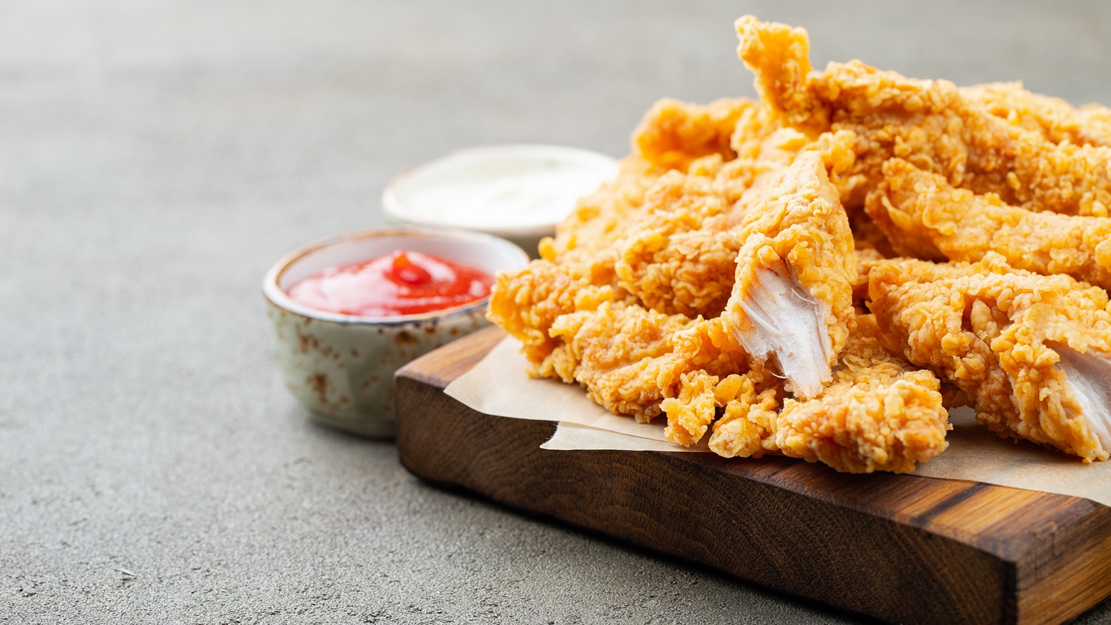 A serving of crispy fried chicken tenders arranged on a wooden board with parchment paper, accompanied by red and white dipping sauces in small bowls.