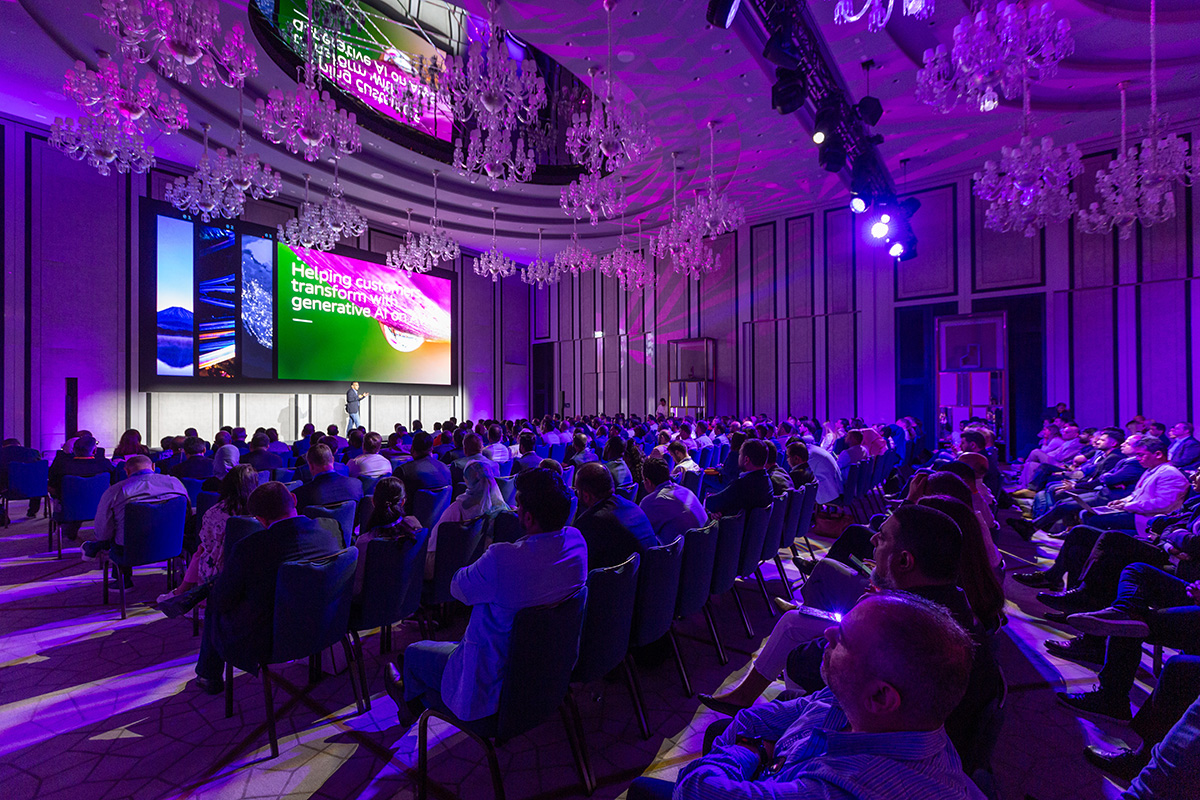 A large audience seated in a purple-lit conference hall, facing a stage with a speaker and a screen displaying a presentation on generative AI.