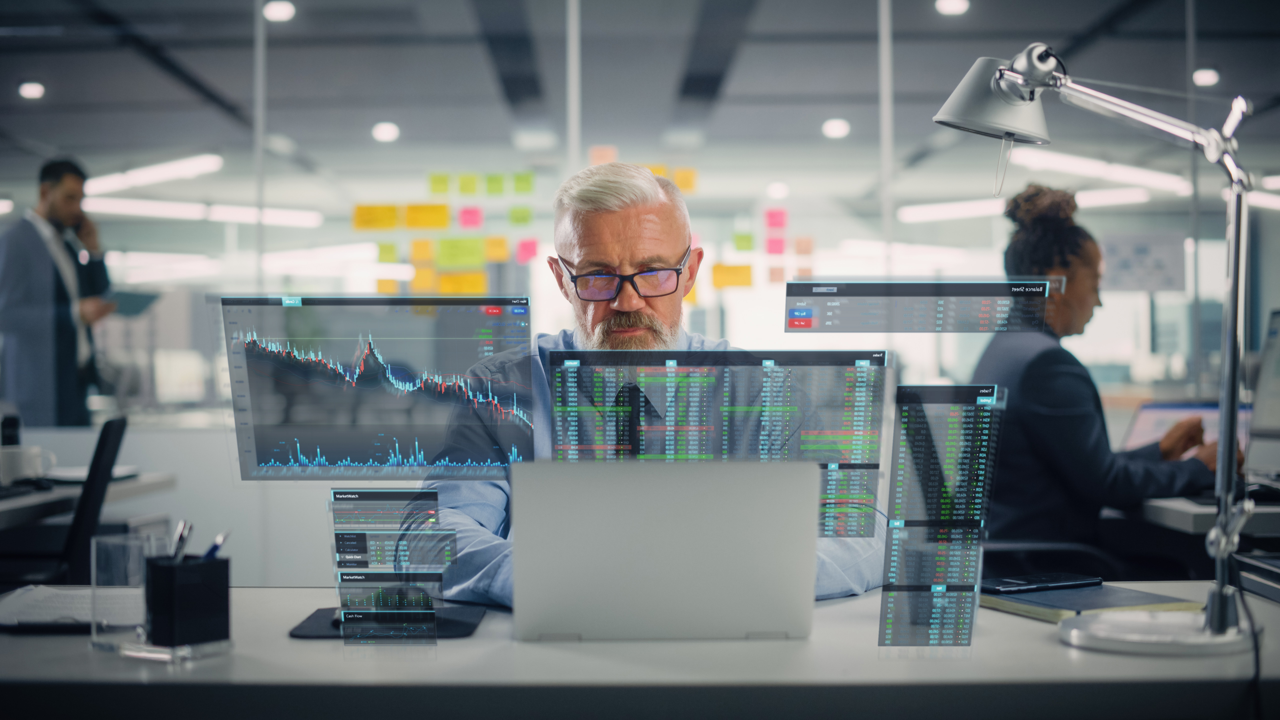 A modern office setting with digital stock market charts and data displayed on transparent screens in front of a laptop, with employees working in the background.