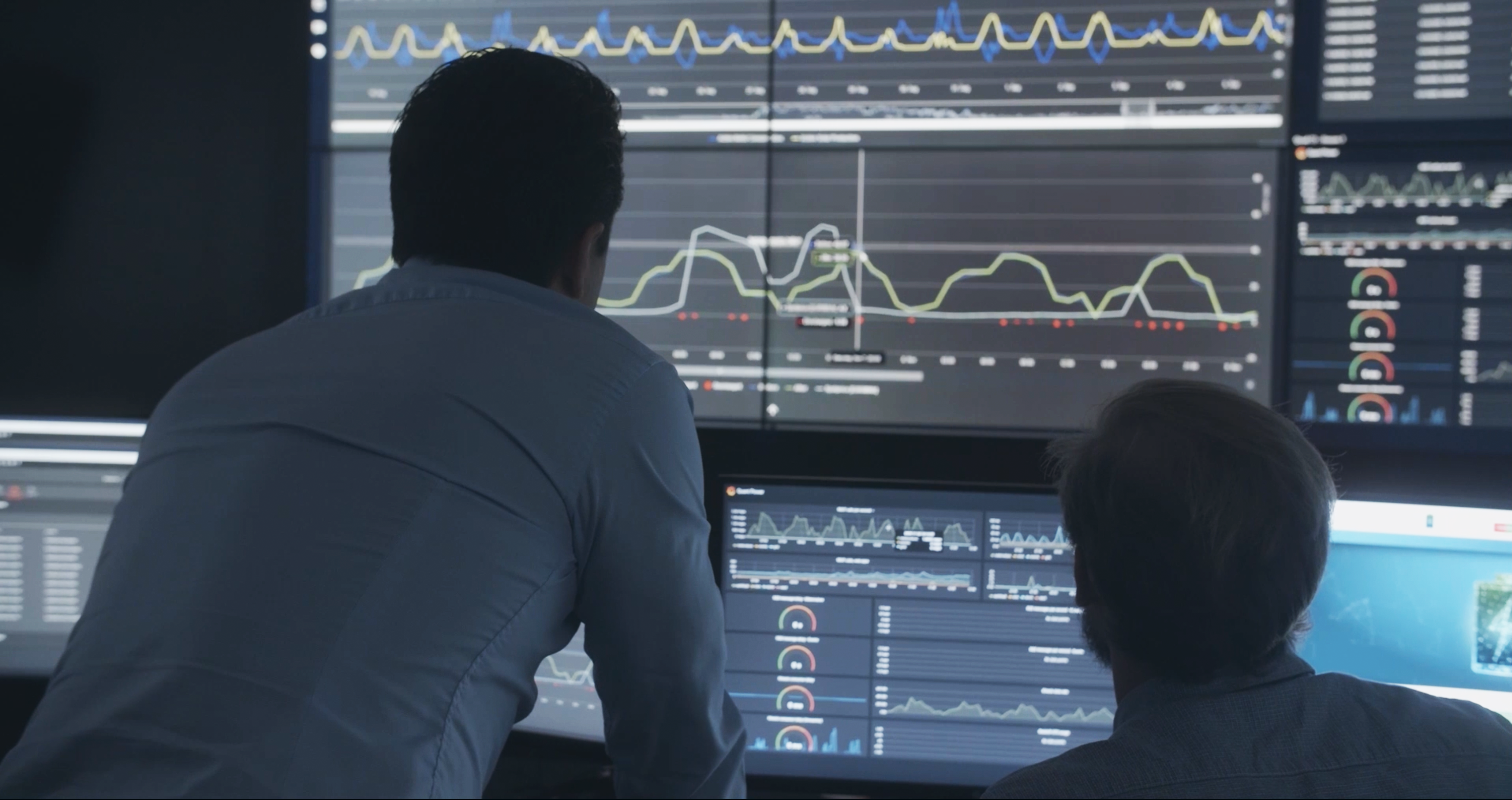 Two people analyzing data visualizations and graphs on multiple large screens in a dark room.