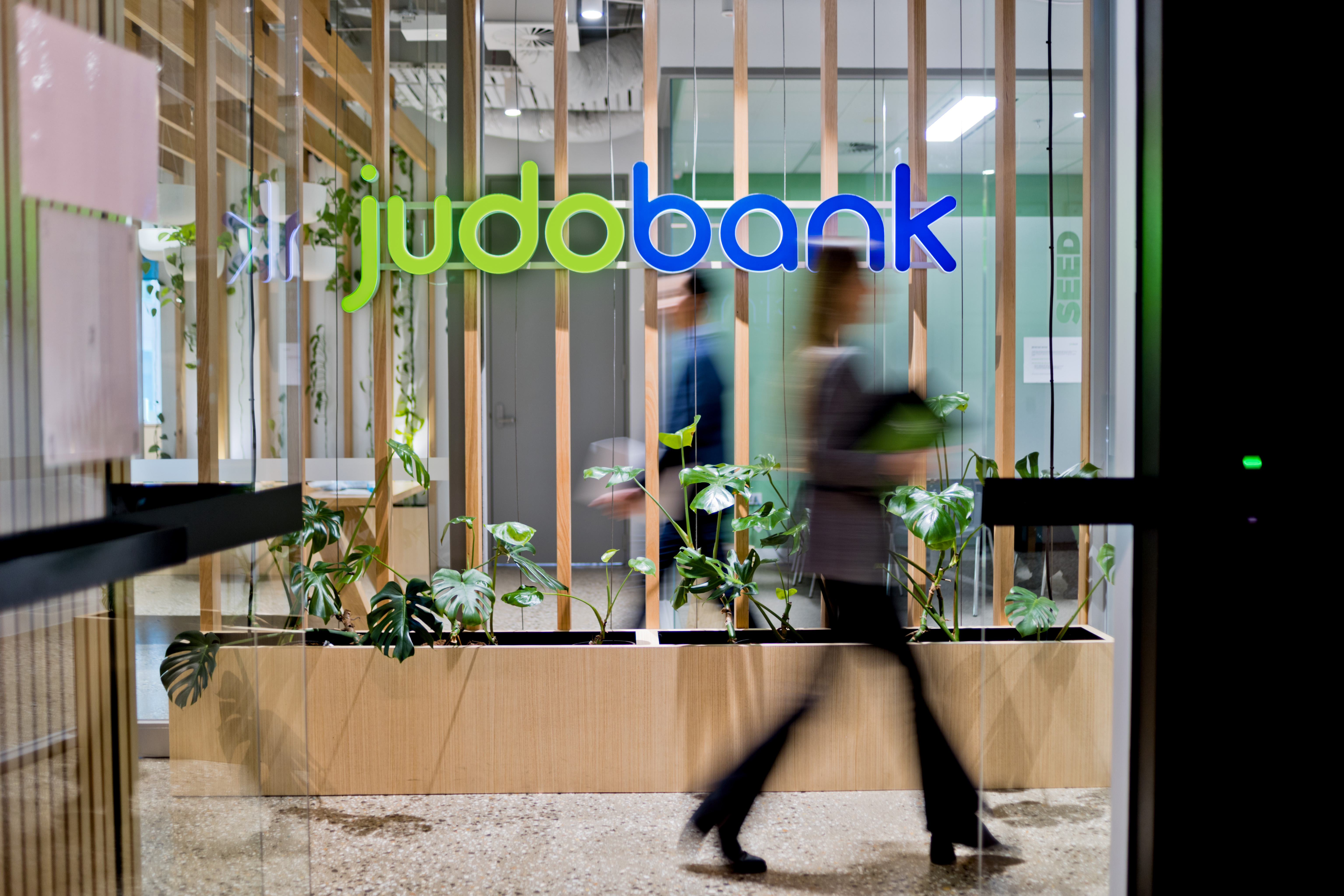 The entrance to a Judo Bank office featuring a modern interior design, with green plants and vertical wooden slats, viewed through glass doors as people walk by.