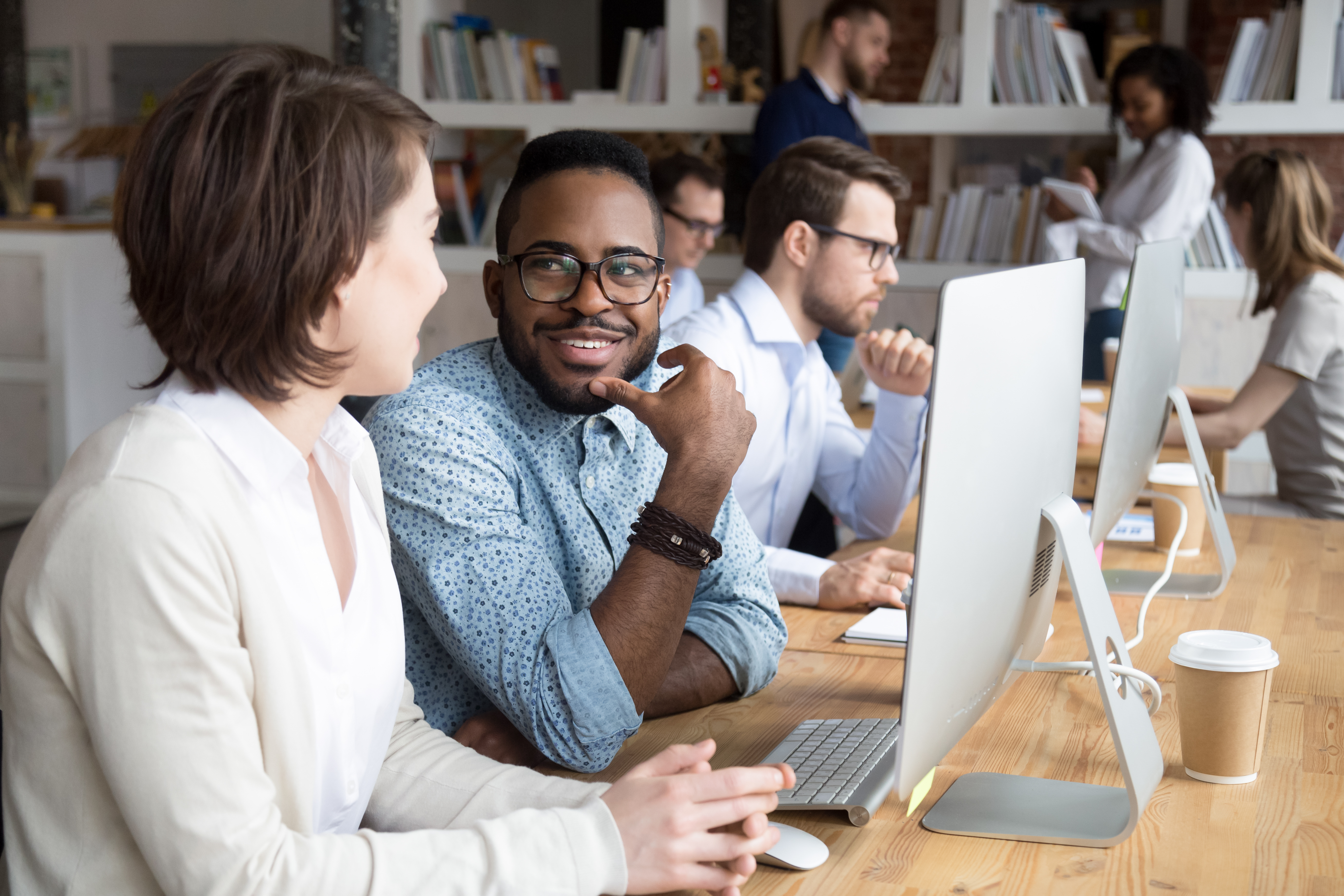 Millennial diverse employees working on contemporary coworking office, on foreground two positive smiling colleagues, mixed race male and caucasian female talking or flirting sitting at desk near pc