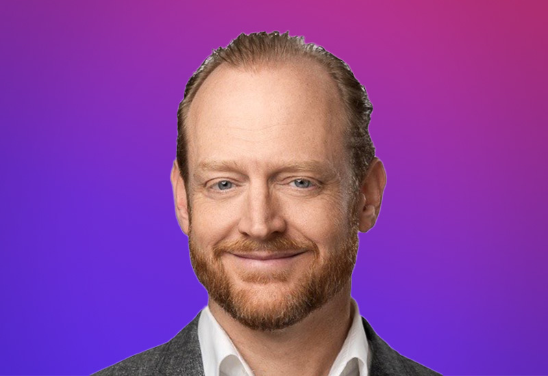 A person wearing a white collared shirt and dark jacket, set against a gradient purple and pink background.