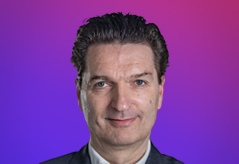 A person with dark, slicked-back hair wearing a suit against a gradient purple and pink background.