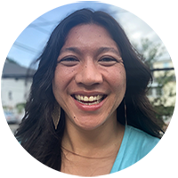 A headshot of a smiling woman with long dark hair, wearing a light blue top, photographed outdoors with a blurred background.