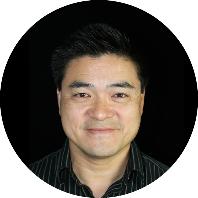 Portrait photograph of an Asian man smiling, wearing a dark striped shirt, against a black background.