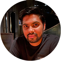 Professional headshot of an individual with short hair and a trimmed beard, seated at a table in a casual setting with dark background.