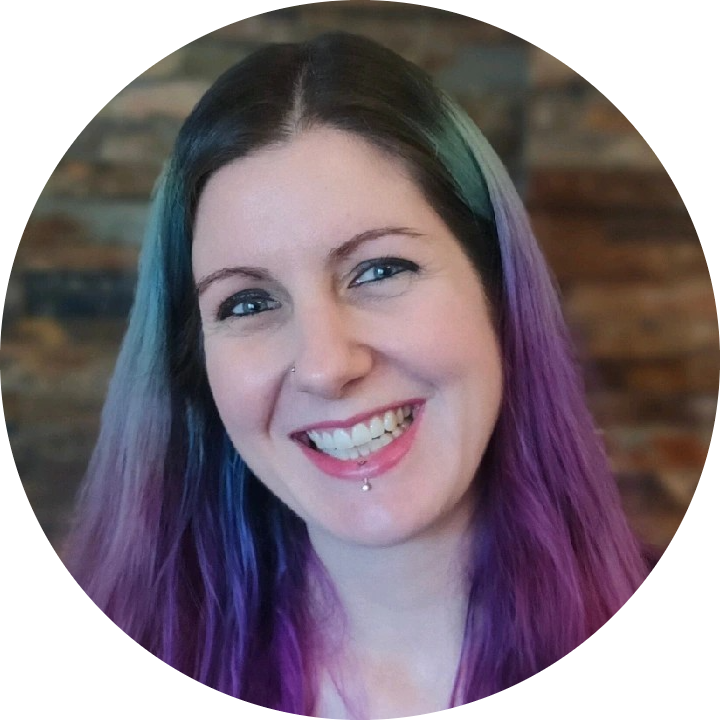 A person with long hair dyed purple and teal, smiling at the camera with a visible lip piercing, in front of a blurred indoor background.