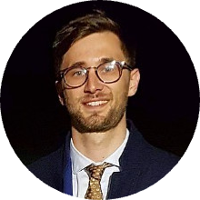 A portrait headshot photo of an individual wearing glasses and business attire, set against a dark background.