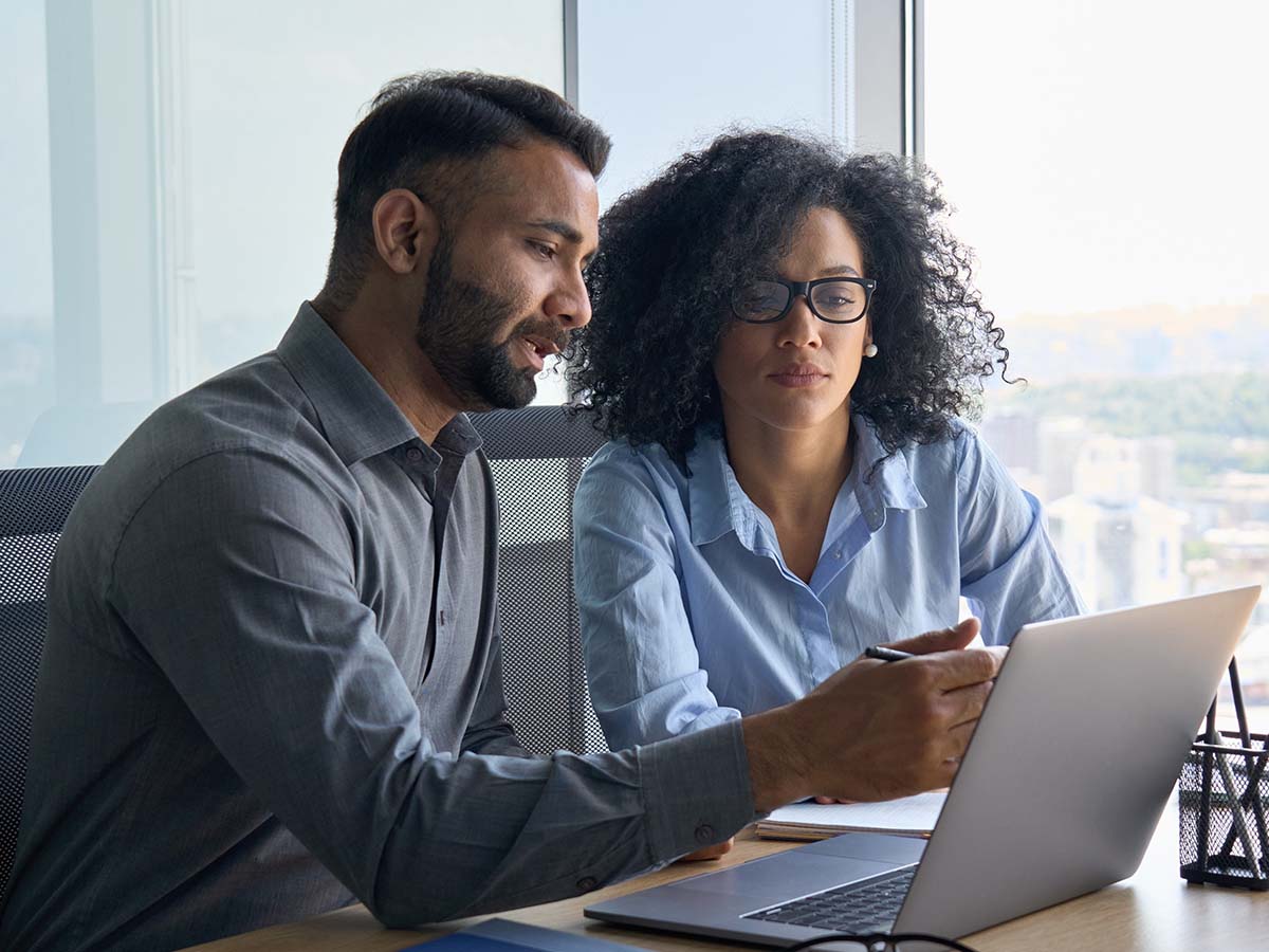Two colleagues working together at a desk, looking at a laptop in a modern office environment with large windows.