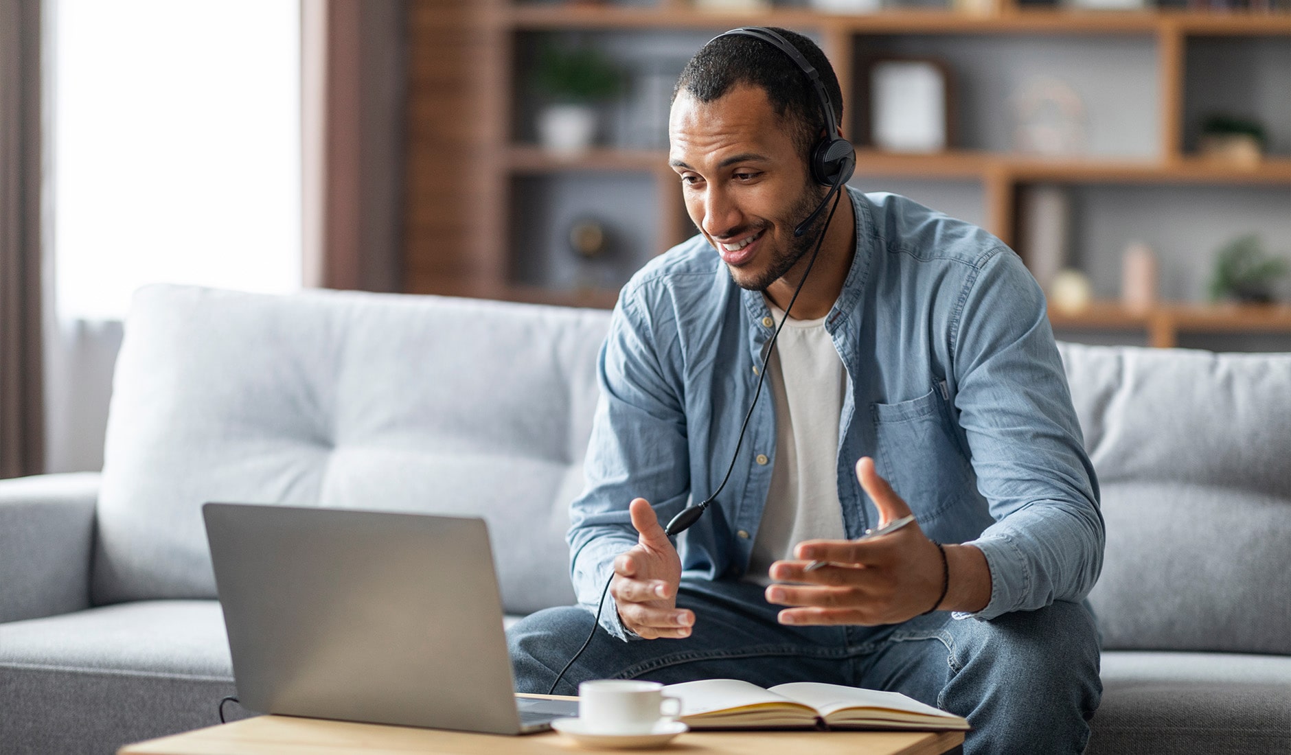 A person wearing headphones gesturing during a video call, seated on a couch with a laptop, notebook, and coffee cup on a table.
