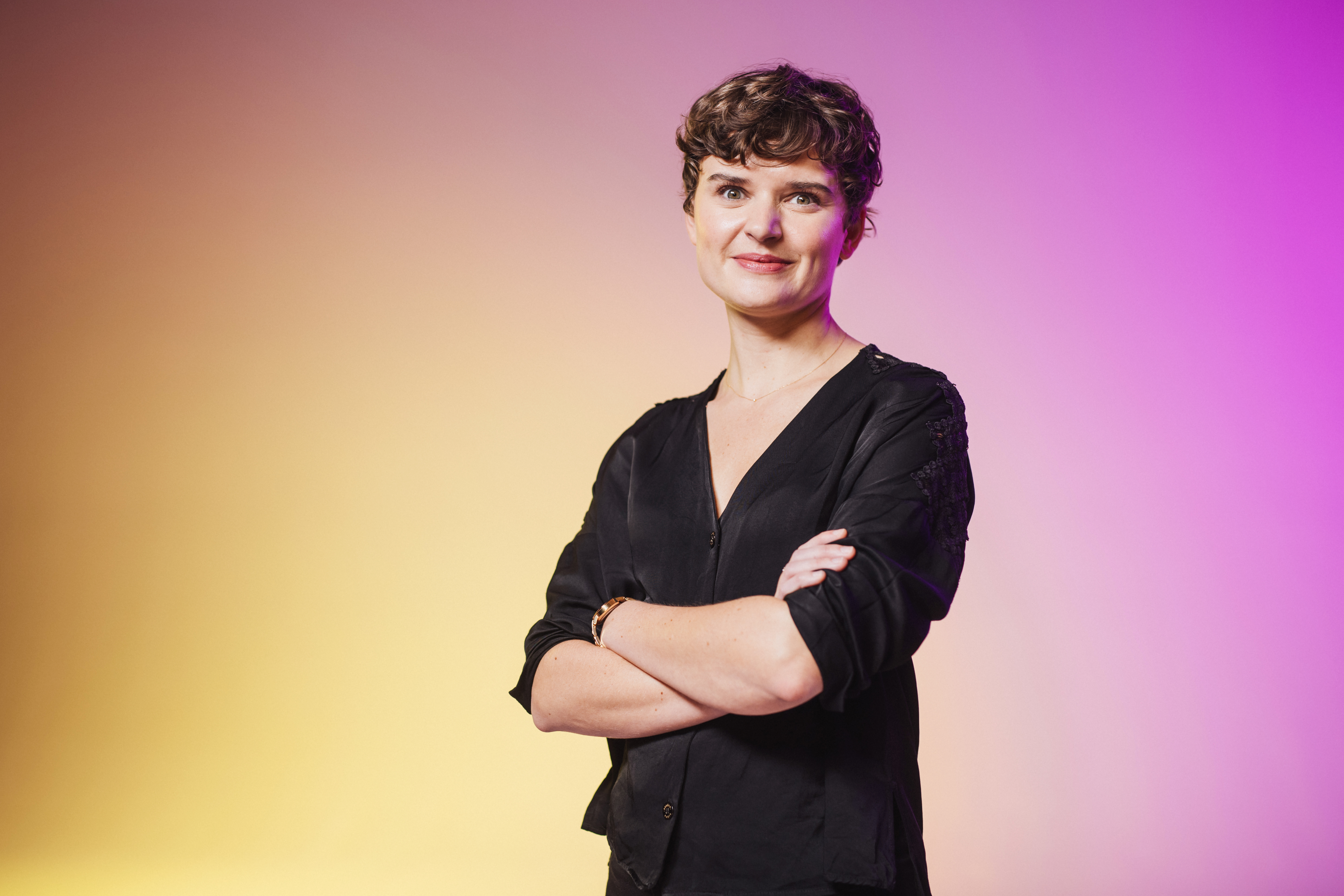 Person wearing a black blouse with arms crossed, standing against a gradient yellow and purple background.