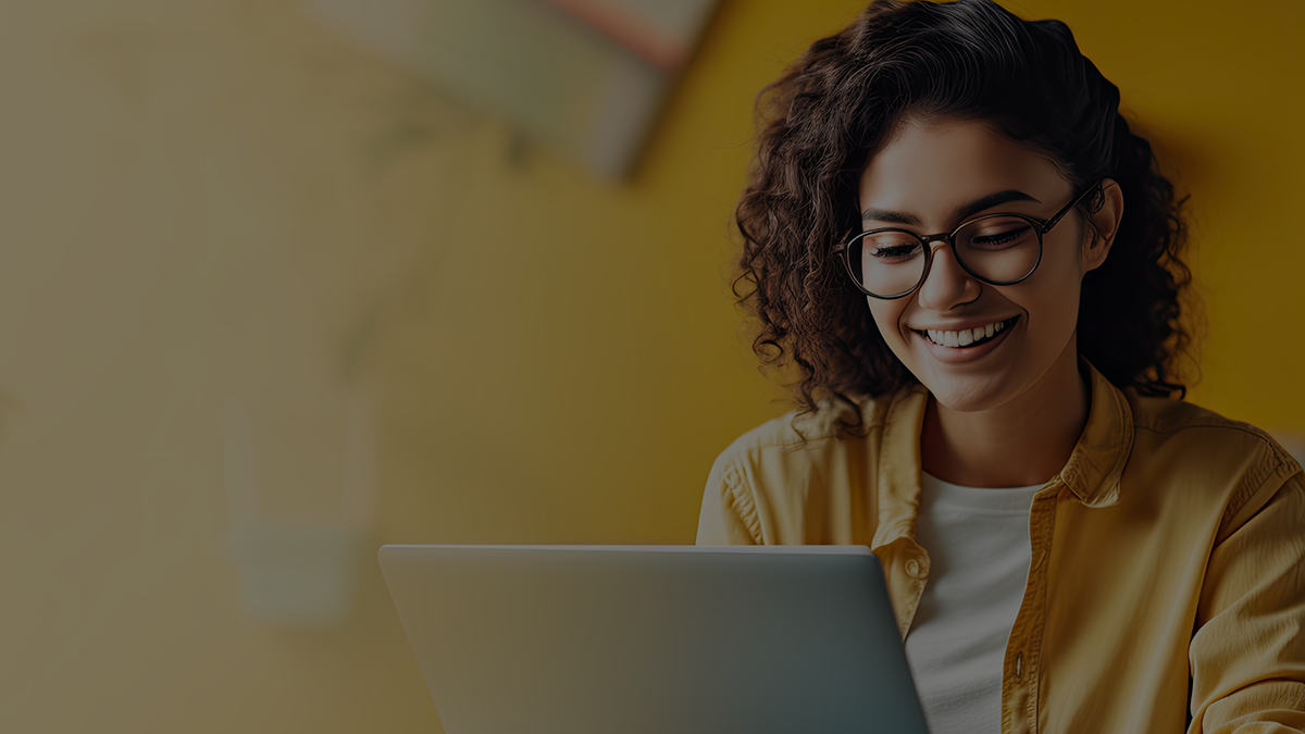 A woman wearing glasses and a yellow shirt is smiling while using a laptop, with a yellow background behind her.