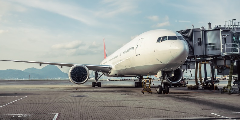 A large commercial airplane is parked at an airport gate, connected to a passenger boarding bridge, with mountains visible in the background.