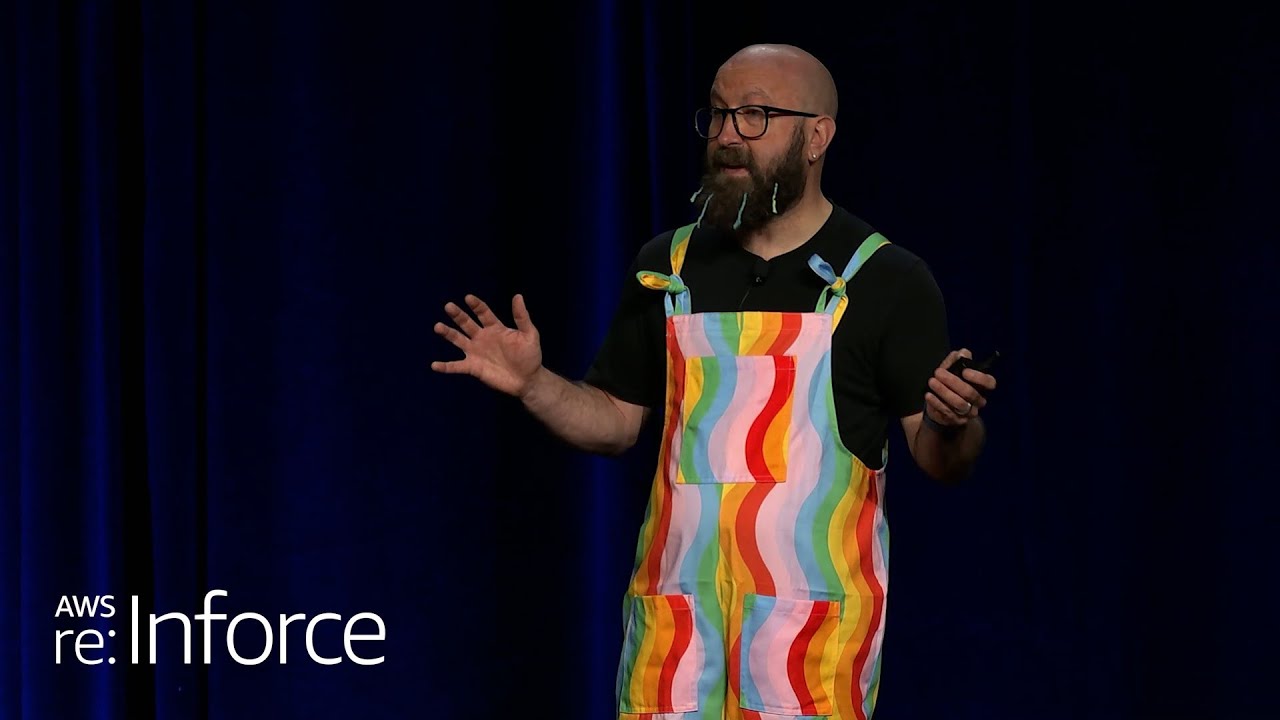 A speaker at AWS re:Inforce presenting on stage, wearing colorful rainbow overalls, with the event logo visible. YouTube thumbnail.