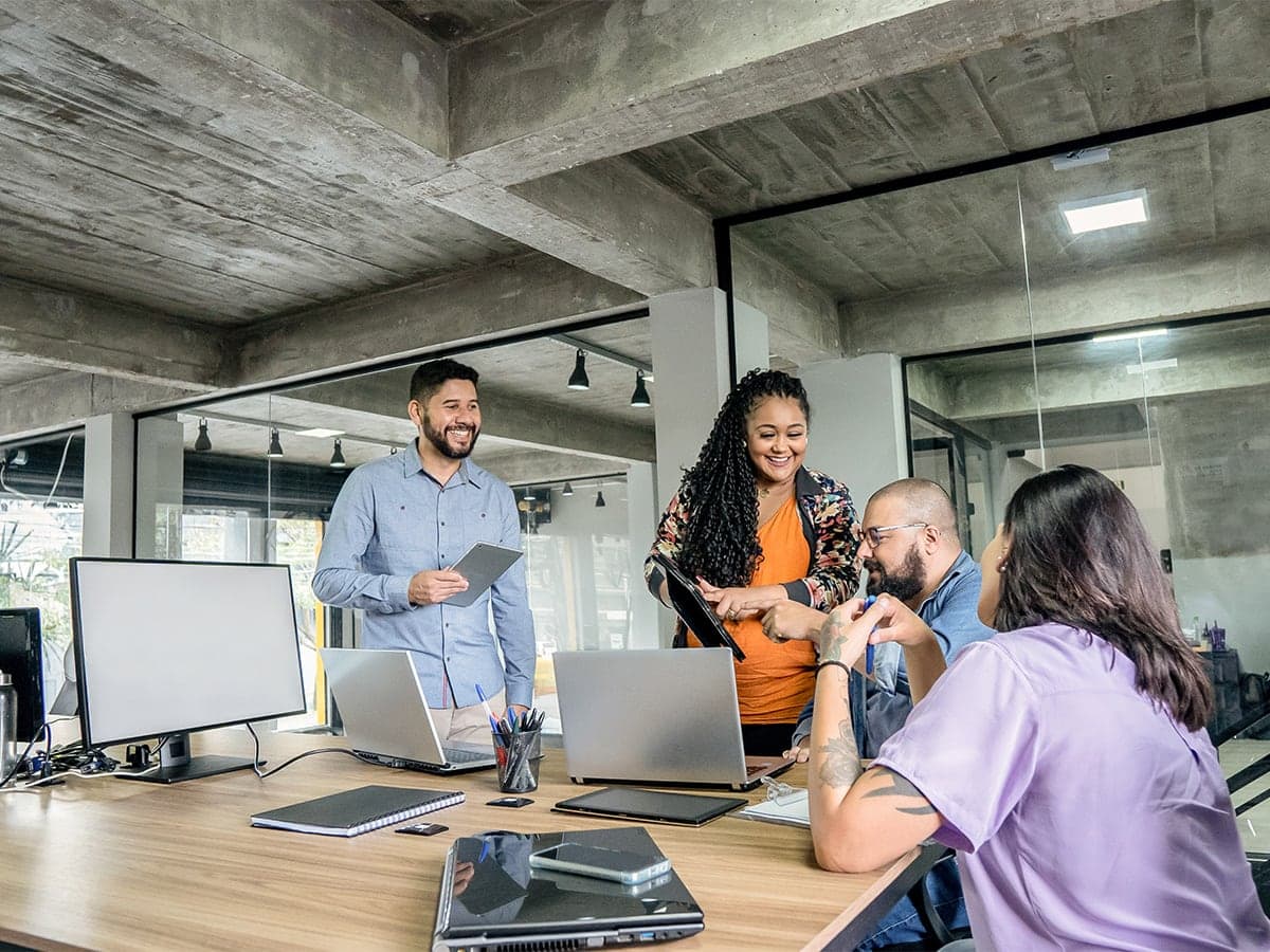 A group of four people collaborating around a desk with laptops and documents in a modern industrial-style office setting.