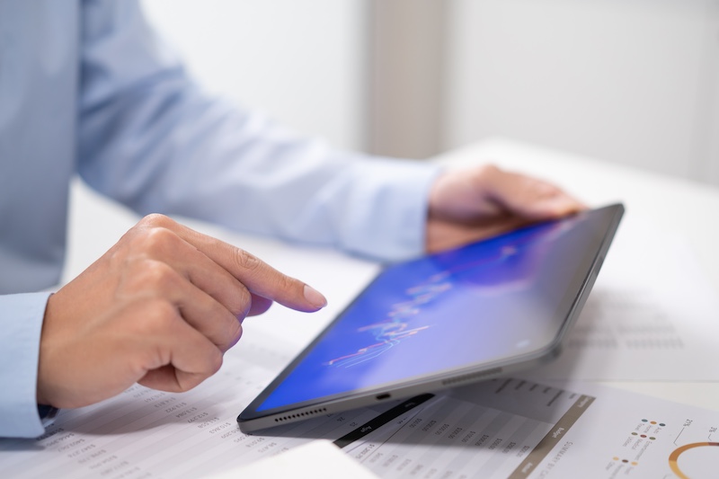 A person in a blue shirt using a tablet device to analyze data charts on a desk, with paper documents and charts visible in the background.