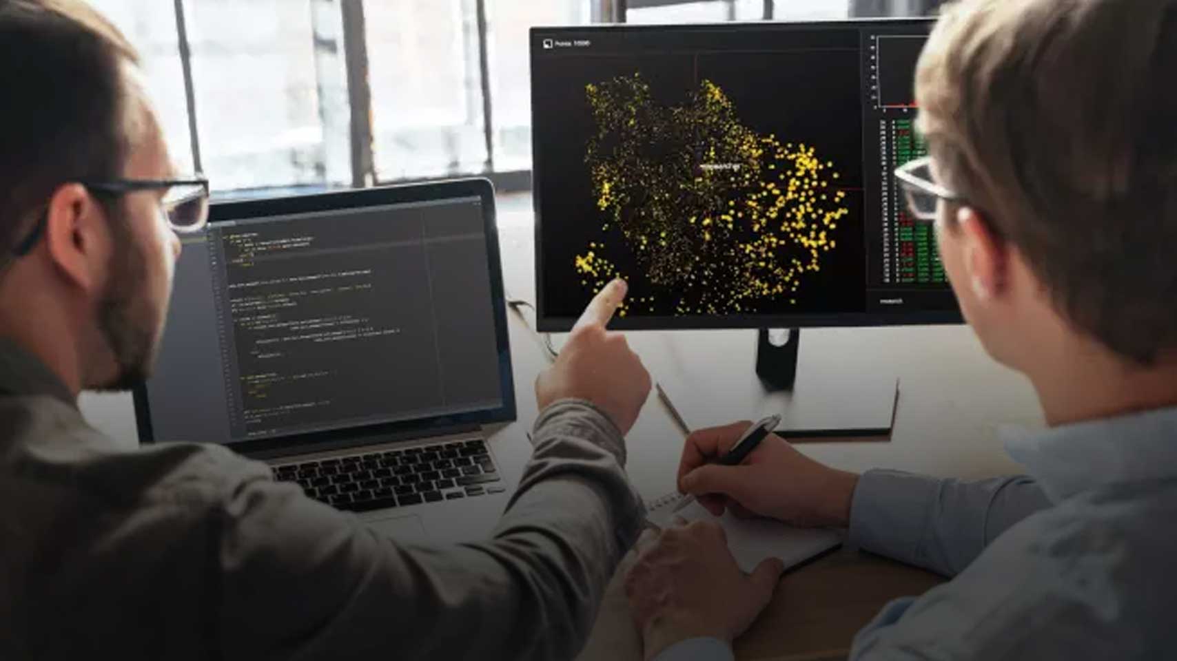 Two people collaborating at a desk, with one working on code displayed on a laptop and the other pointing to a data visualization of yellow data points on a computer monitor, representing artificial intelligence or machine learning analysis.