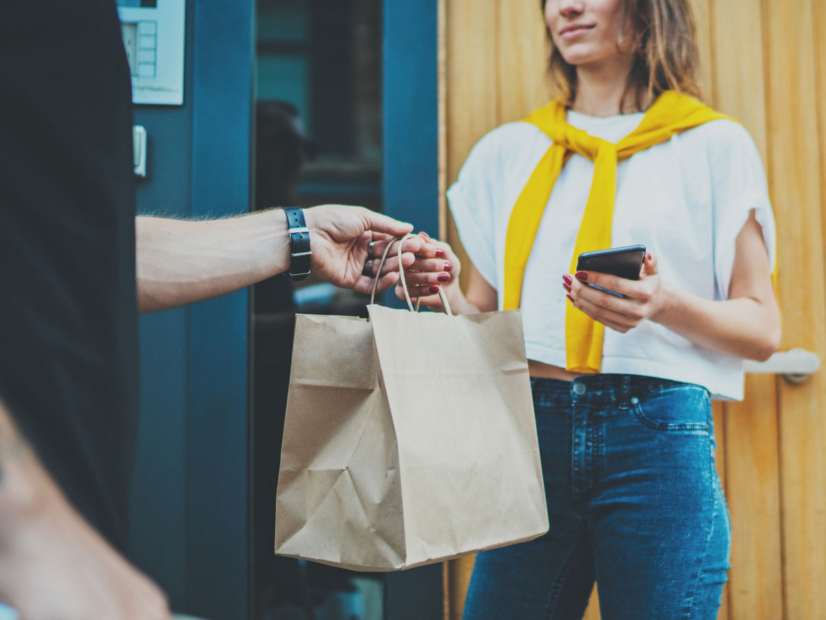 A person receives a paper bag likely containing food or groceries from a delivery worker at their doorstep, illustrating contactless delivery using a mobile phone.