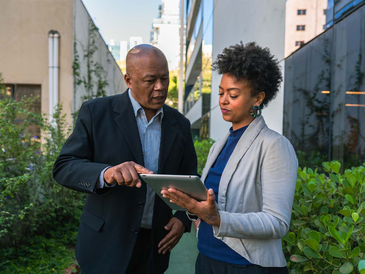 Two coworkers are standing outdoors in a modern business environment, collaborating while looking at and pointing to a tablet device.