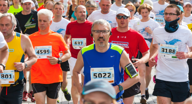 A group of marathon runners competing in a race, each wearing numbered race bibs and athletic attire. The participants are running closely together outdoors on a bright day.
