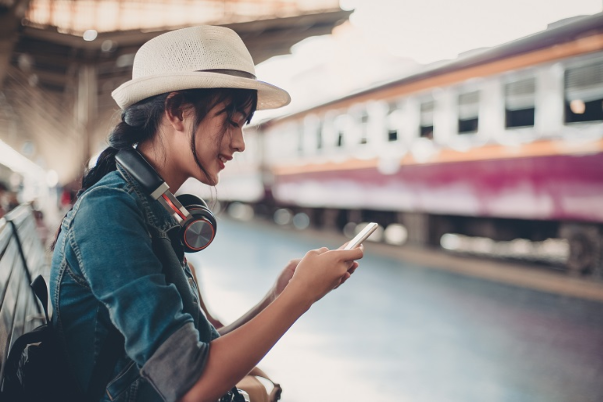 A young traveler wearing headphones and a hat uses her smartphone while waiting at a train station, with a train in the background.